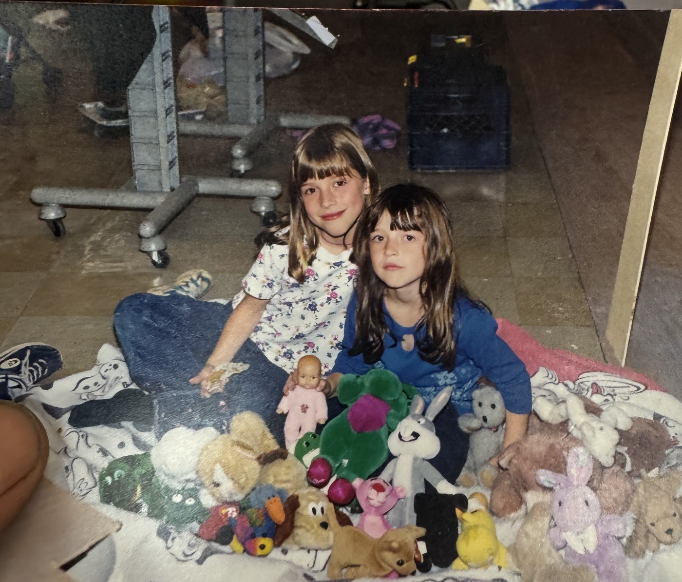 Two girls sitting on a blanket on the sales floor surrounded by stuffed toys, with rolling garment racks behind them