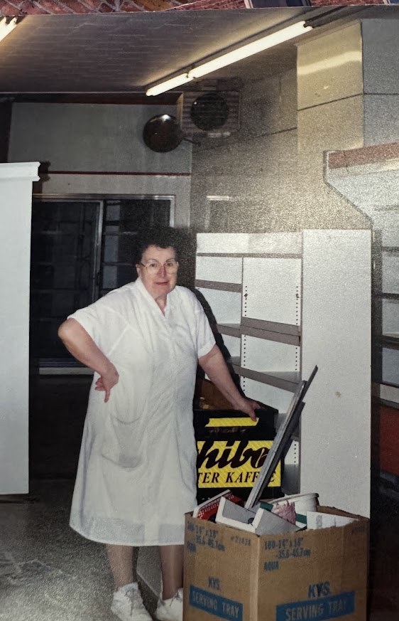A woman in a white work coat standing among stripped shelving and packing boxes at the end of Link's Delicatessen