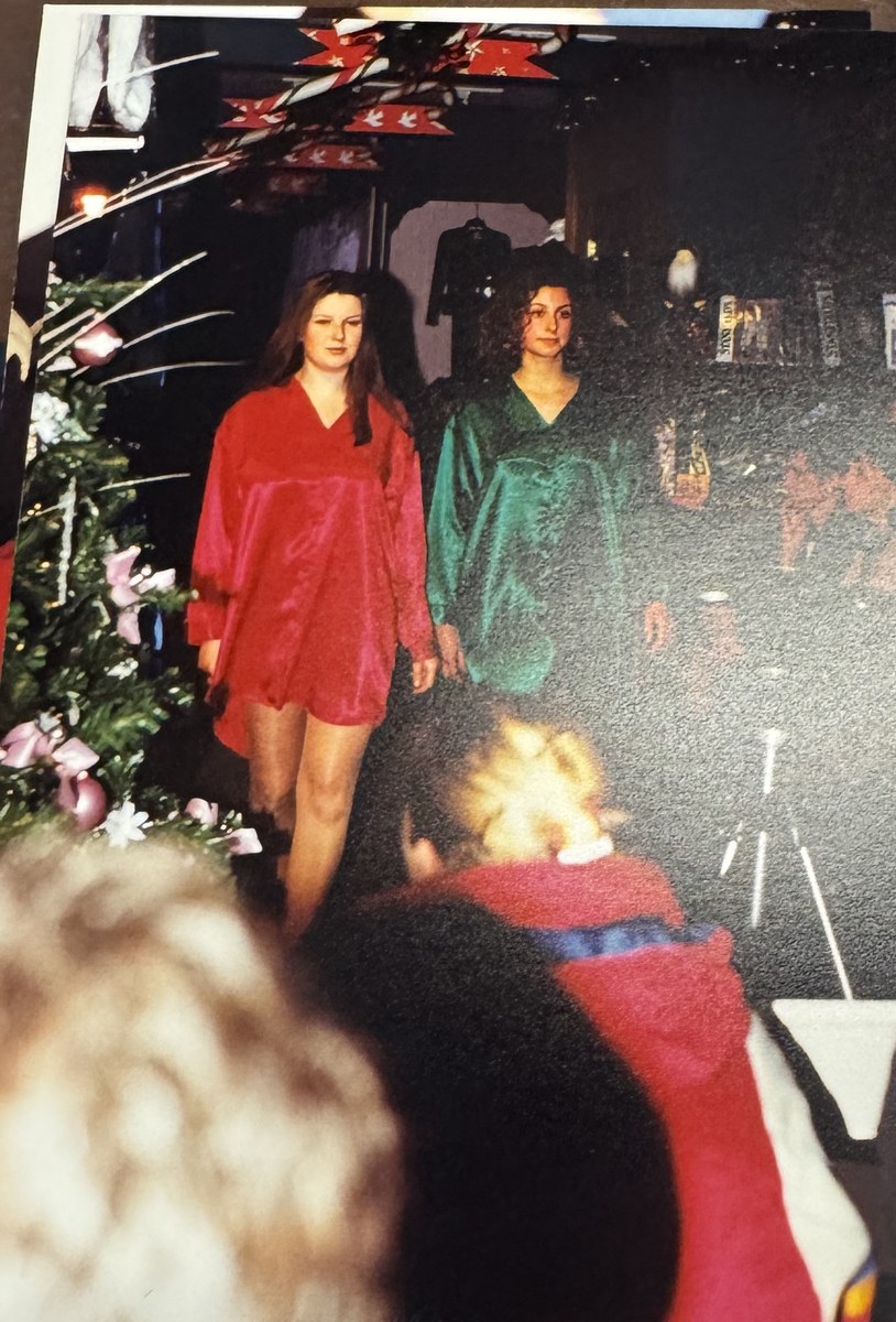Two models walking through the shop past a decorated tree during an in-store fashion show, with a seated audience in the foreground