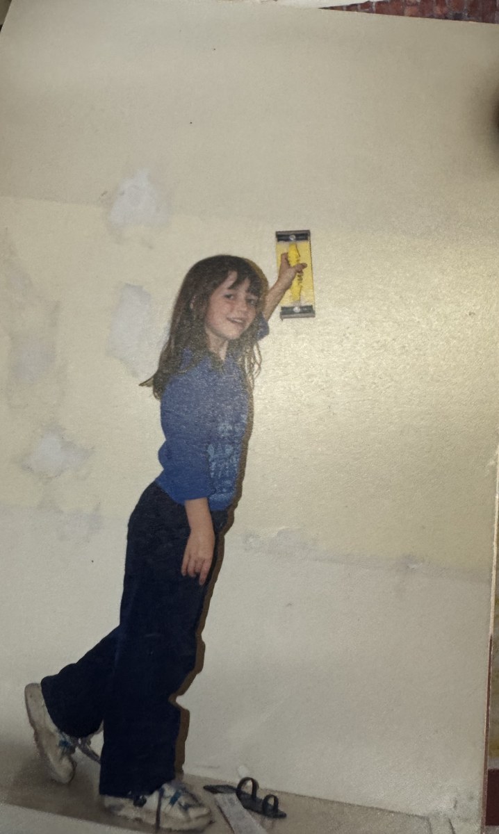 A young girl on the shop floor holding a scraper up against a stripped and patched wall during a renovation